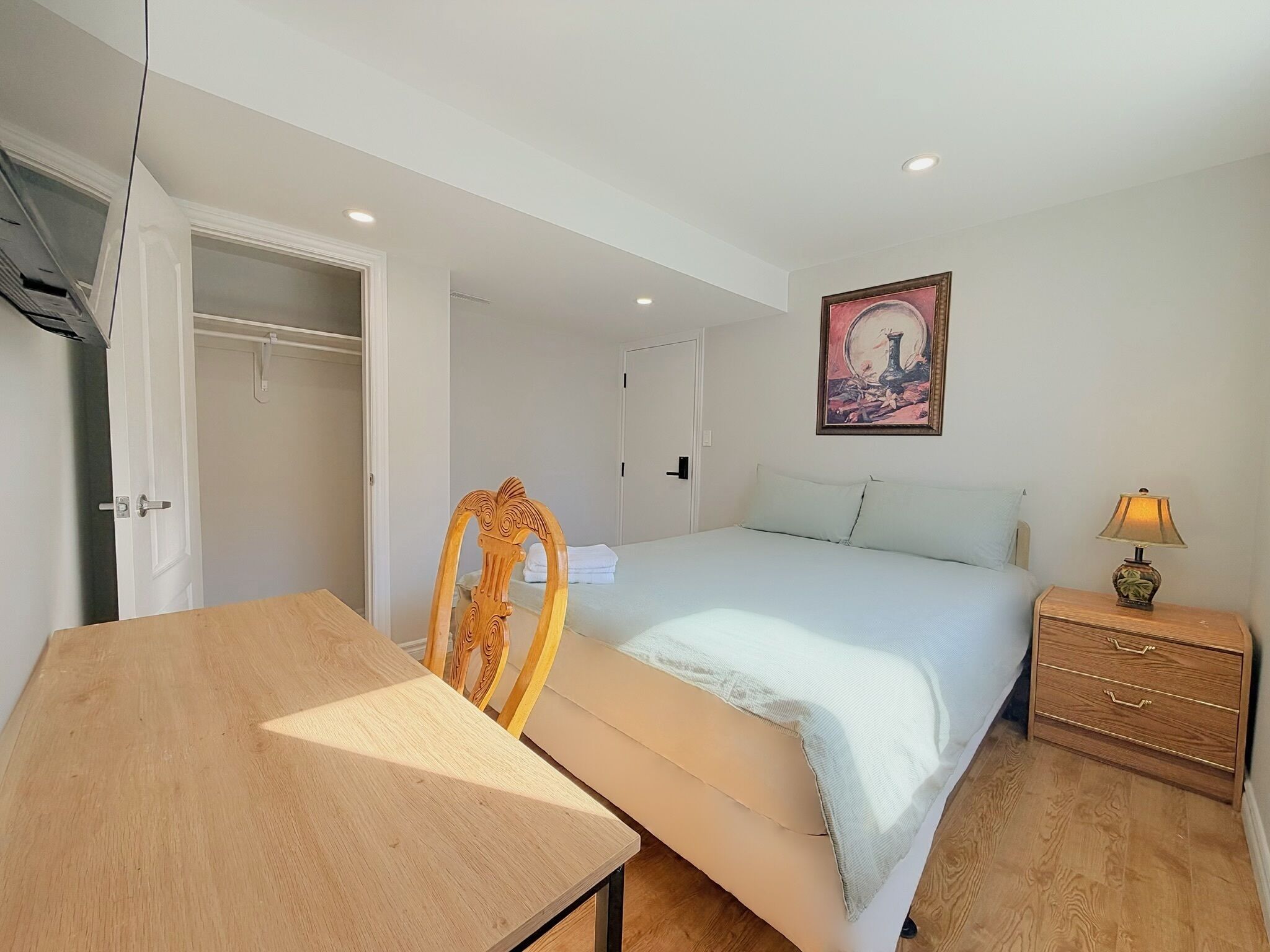 Une chambre à coucher avec un lit, une chaise en bois, une table de chevet avec une lampe et un tableau accroché au mur.