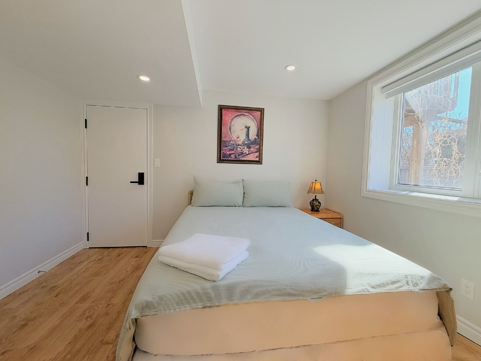 A bedroom with a bed, a door, a window, and a painting on the wall.