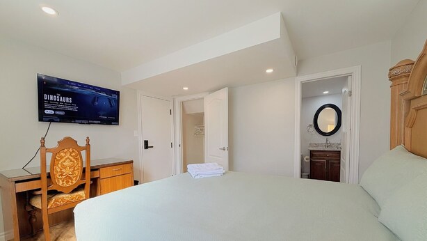 Un dormitorio con cama, televisor montado en la pared, una silla de madera, escritorio y puerta de baño.