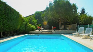 A rectangular swimming pool with a clear blue water surface, surrounded by a concrete deck. There are several white lounge chairs and a couple of potted plants with red flowers.