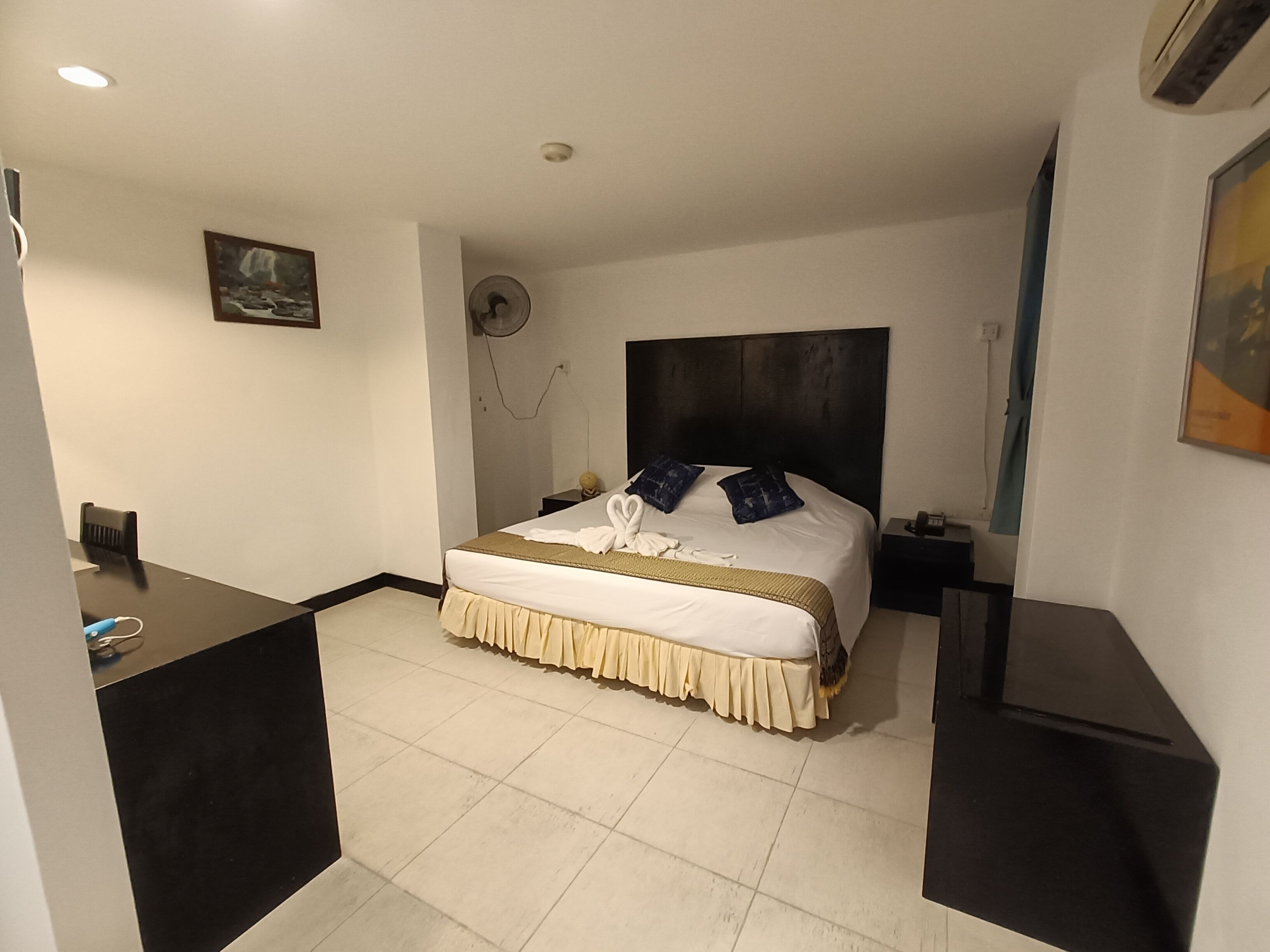 A hotel room with a bed, bedside tables, a desk, and a fan.