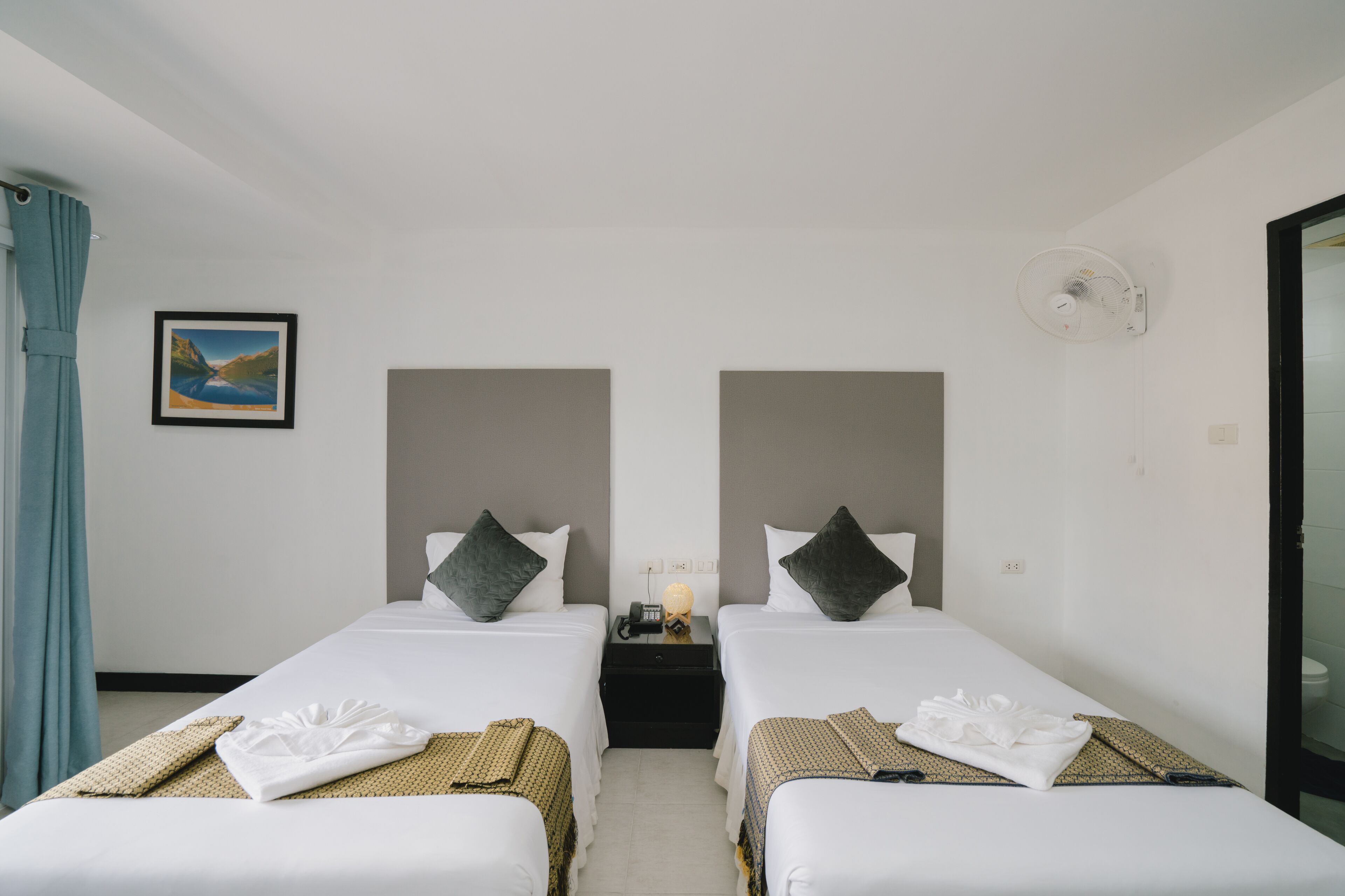 Une chambre d’hôtel avec deux lits, une table de chevet, un ventilateur fixé au mur et un tableau encadré.