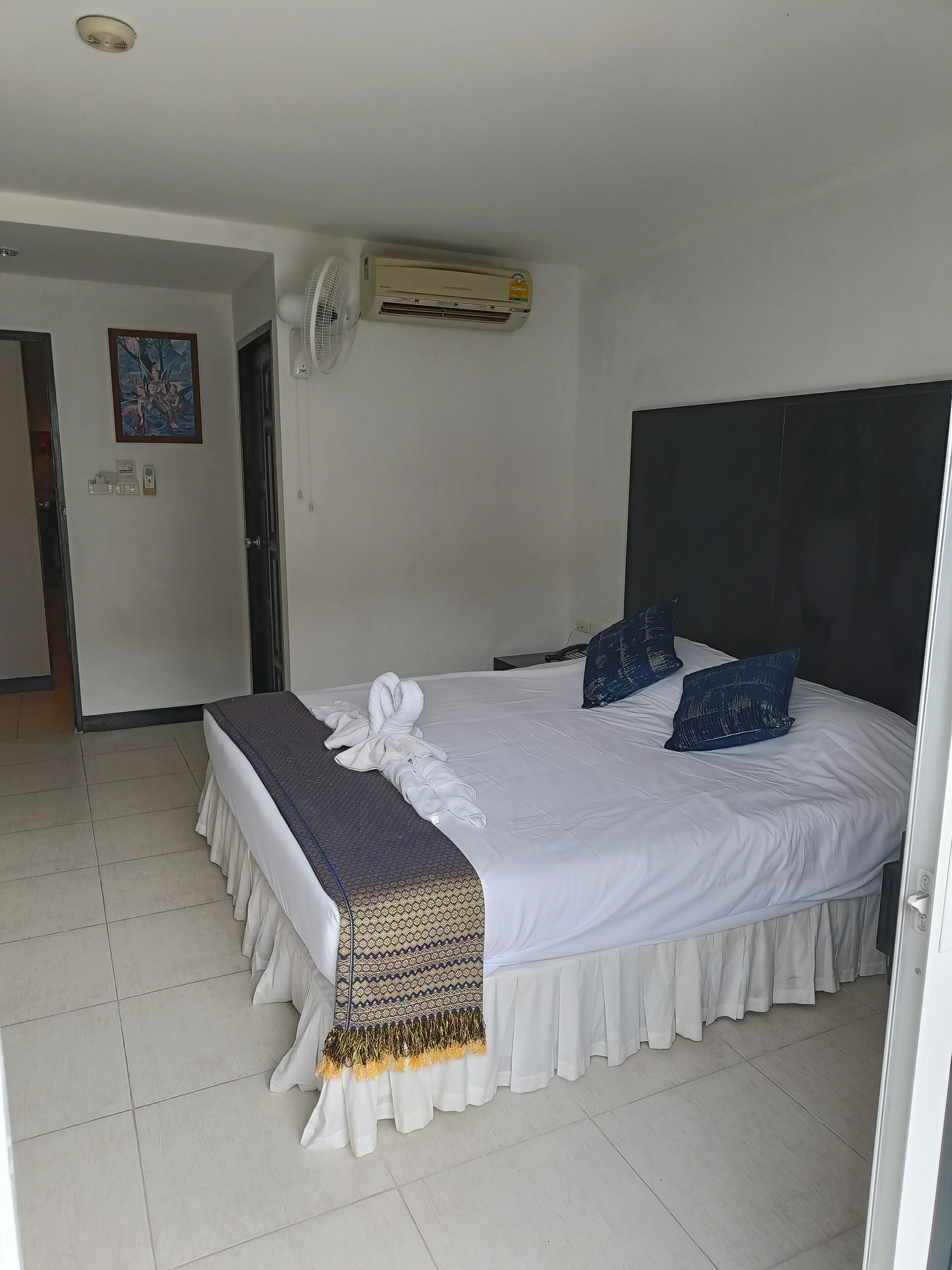 A hotel room with a bed, a wall-mounted air conditioner, a painting, and a door.