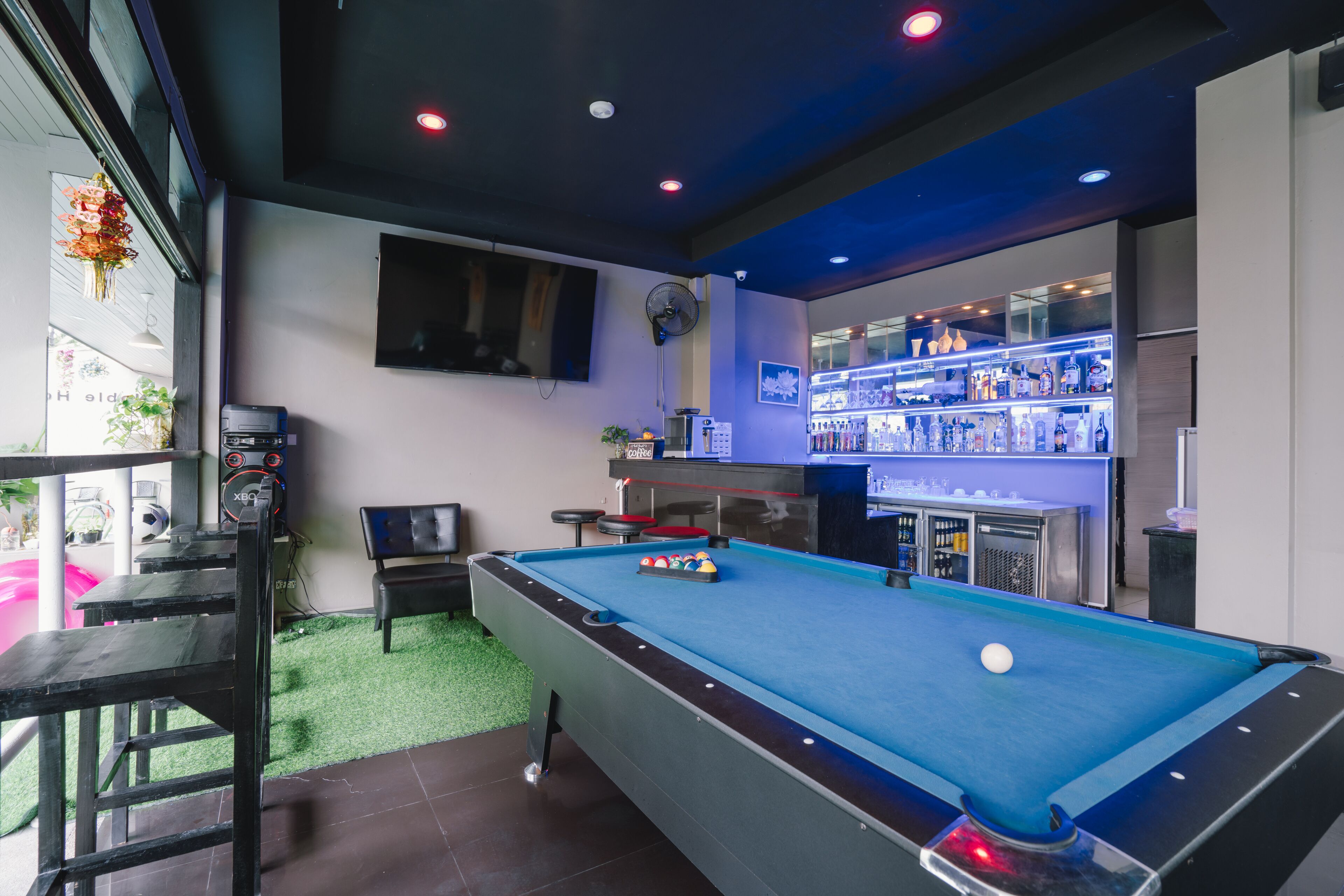 Una mesa de billar con superficie de fieltro azul, una zona de bar con estanterías iluminadas y un área de descanso con un banco y una mesa.