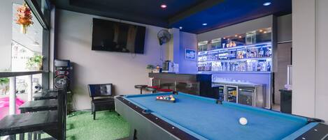 Una mesa de billar con superficie de fieltro azul, una zona de bar con estanterías iluminadas y un área de descanso con un banco y una mesa.