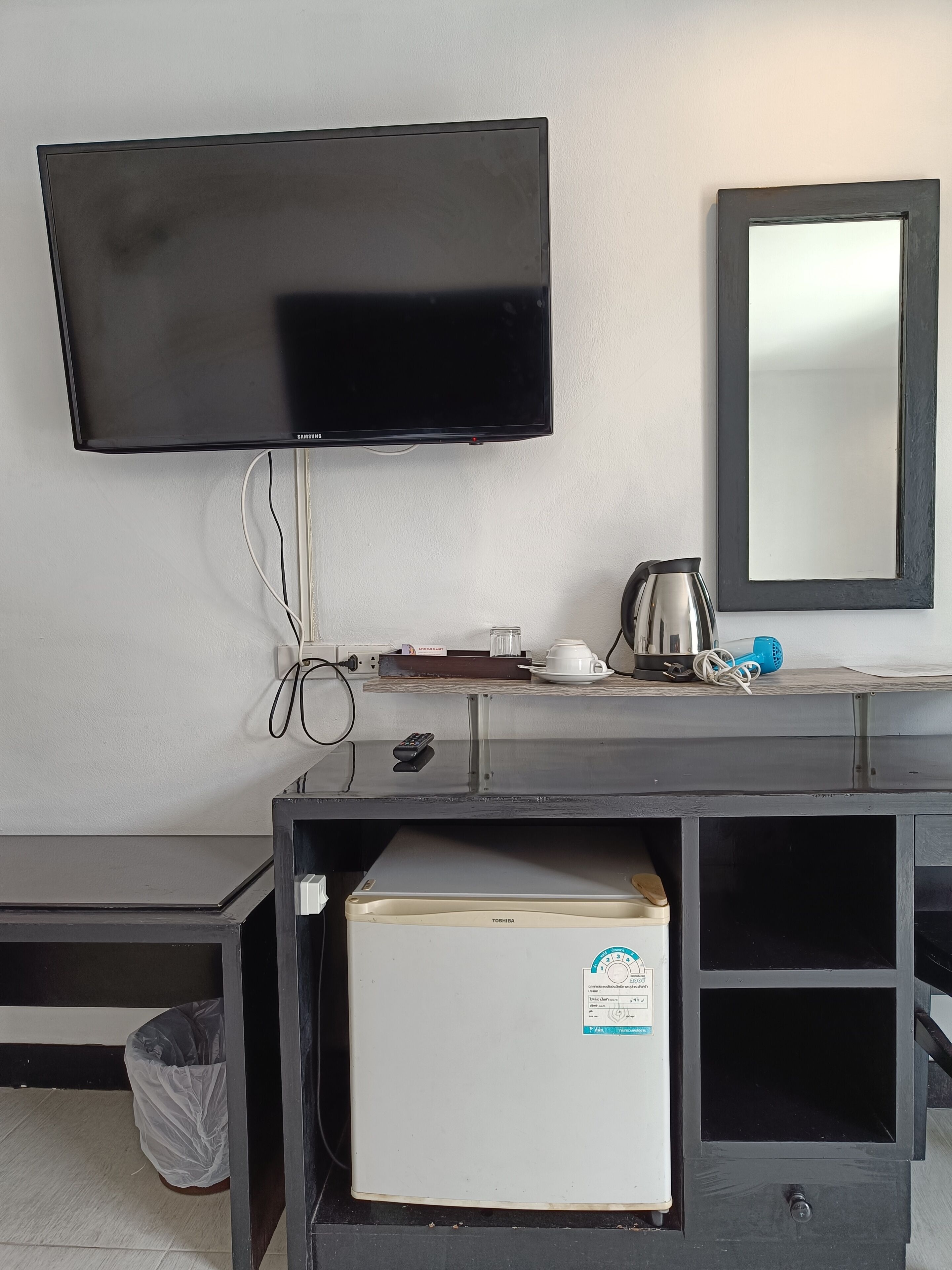 A room with a flat-screen TV mounted on the wall, a small table with a kettle, a cup, and a remote control. There is a refrigerator with a sticker on it, and a trash bin is visible.