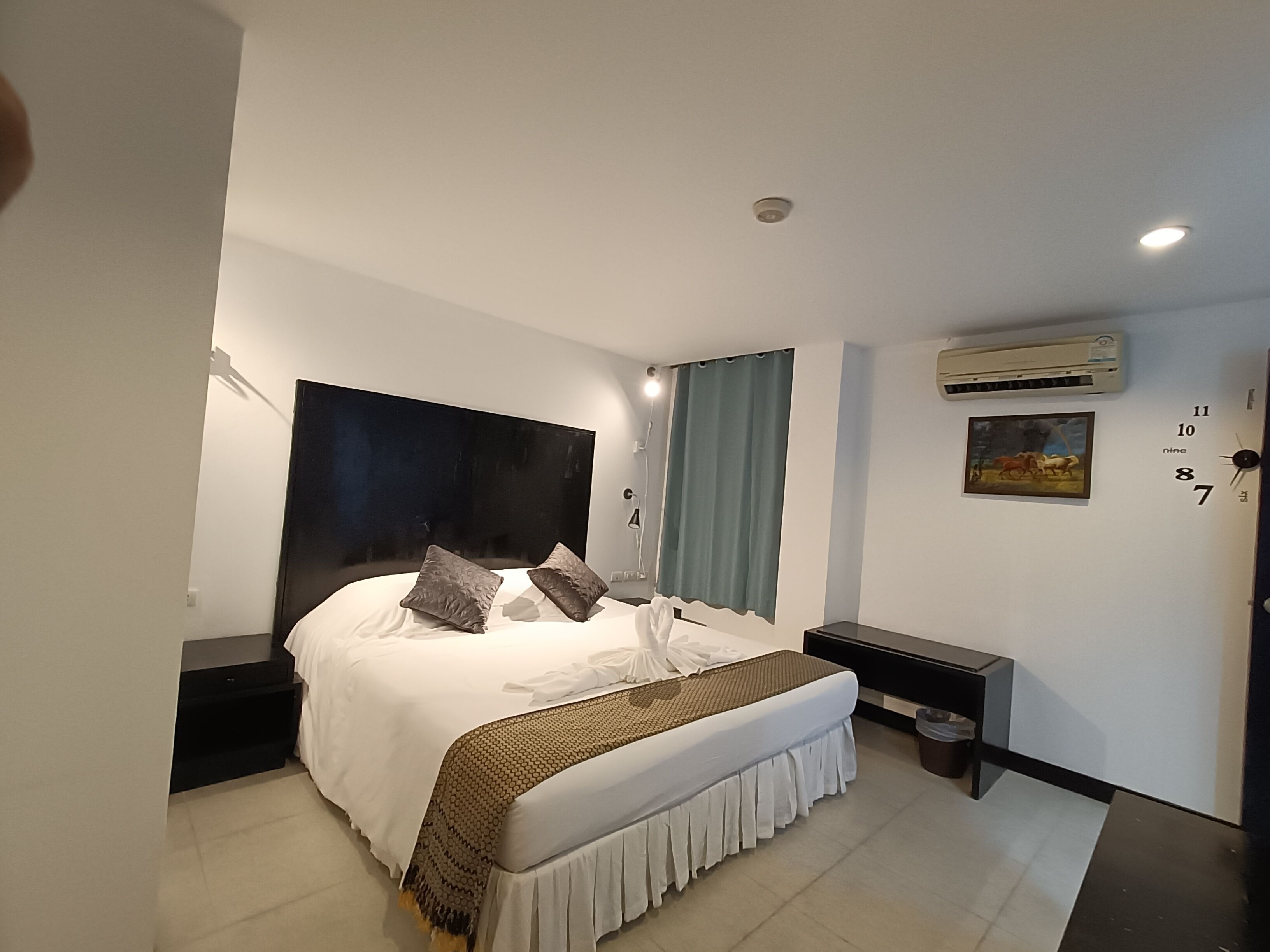 A modern bedroom with a large bed, bedside tables, an air conditioning unit, and a wall clock.