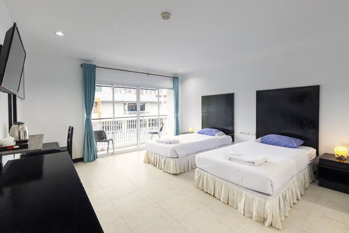 Une chambre d’hôtel avec deux lits, une télévision, un bureau avec une bouilloire et un balcon donnant sur les bâtiments.