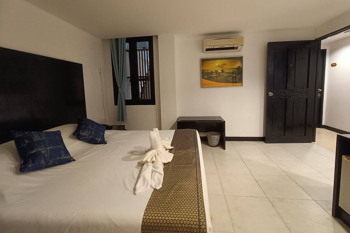 Une chambre d’hôtel avec un lit, une petite table de chevet, une poubelle, un tableau au mur et une porte.