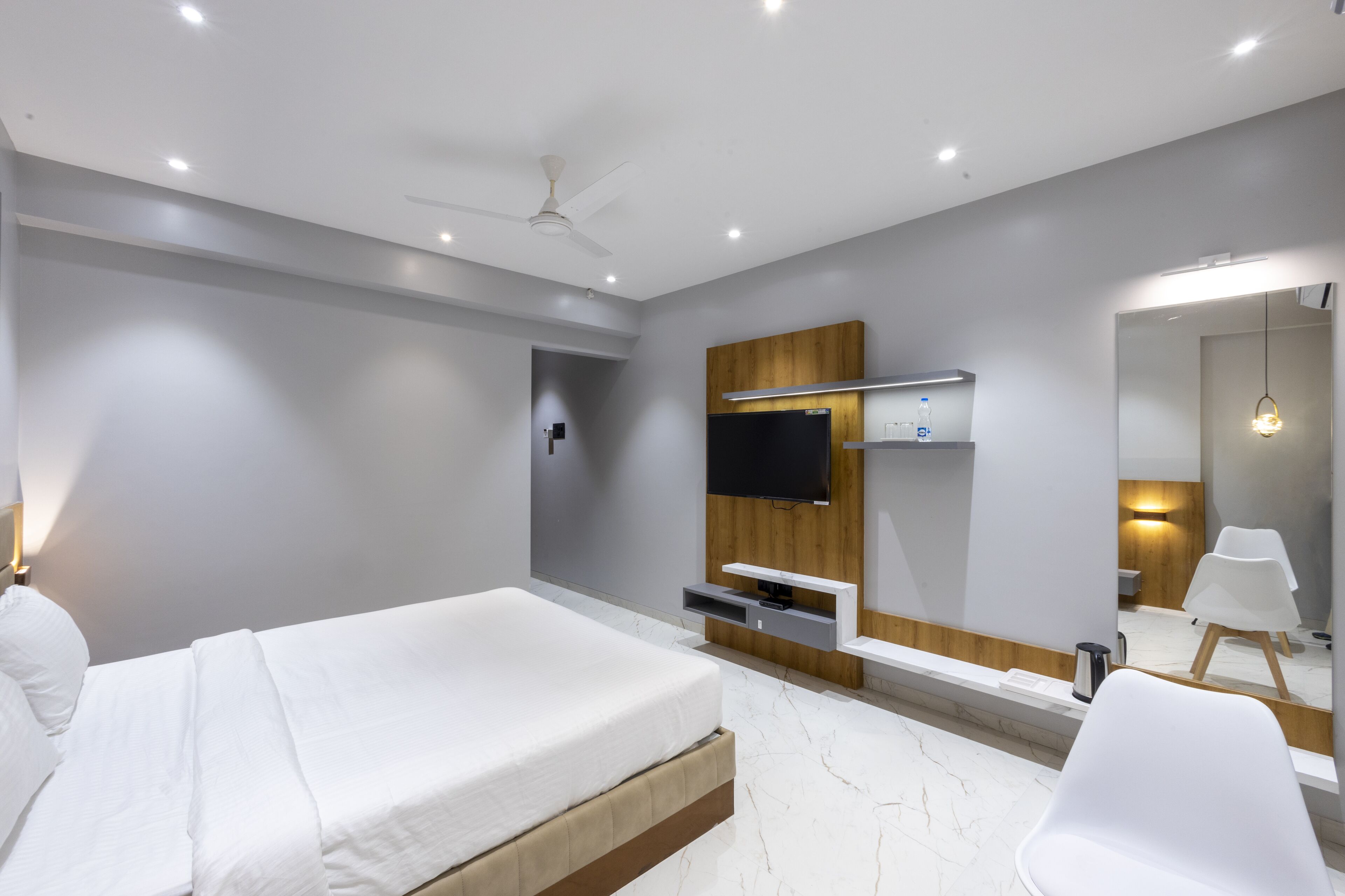 A modern hotel room with a bed, a TV mounted on a wooden panel, a mirror, and a white chair.