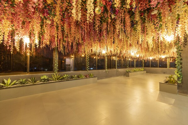 An indoor space with hanging floral decorations, large windows, and potted plants along the walls. - Hotel Opulence (Chhatrapati Sambhajinagar)