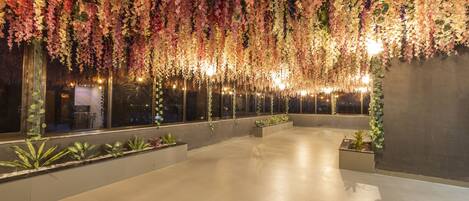 A spacious indoor area with a high ceiling adorned with hanging floral arrangements, large windows, and potted plants along the walls.