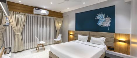 A modern hotel room with a large bed, a ceiling fan, an air conditioning unit, a decorative wall piece, and a seating area with chairs.