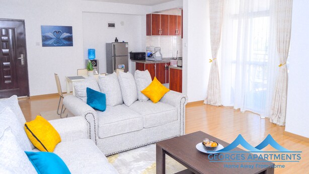 A living room with a grey sofa, blue and yellow cushions, a wooden coffee table, and a kitchen in the background. - George's Fully Furnished 2 Bedroom Apartment (Nairobi)