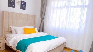 A bedroom with a bed, a nightstand, a window with curtains, and a logo on the wall. - George's Fully Furnished 2 Bedroom Apartment (Nairobi)