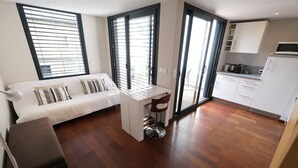 Ein modernes Wohnzimmer mit einer Couch, einem kleinen Tisch und einem Barhocker. Es gibt eine Küchenzeile mit Schränken und einer Mikrowelle.