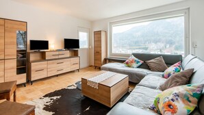 A modern living room with a sofa, a coffee table, a TV, and a large window with a mountain view. - Holiday Home Feldkirch Panoramic Views (Feldkirch)