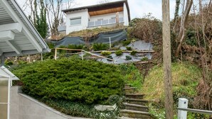A modern house with a sloped roof, a staircase leading to a deck, and a garden area with various plants. - Holiday Home Feldkirch Panoramic Views (Feldkirch)