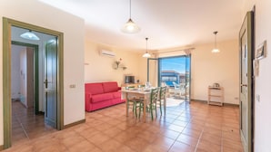 A spacious living area with a red sofa, a dining table with chairs, and a view through a glass door. - Residence I Mirti Bianchi (Santa Teresa di Gallura)