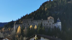 Un edificio parecido a un castillo con múltiples torres y un techo cónico destacado, rodeado de un bosque denso y colinas.