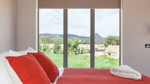 Un dormitorio con cama, almohadas rojas y blancas, una colcha roja y vistas a un paisaje con vegetación y un edificio.