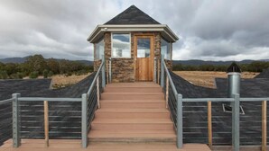 Una pequeña estructura de piedra y madera con puerta de madera y ventanas amplias, a la que se accede por una escalera de metal.