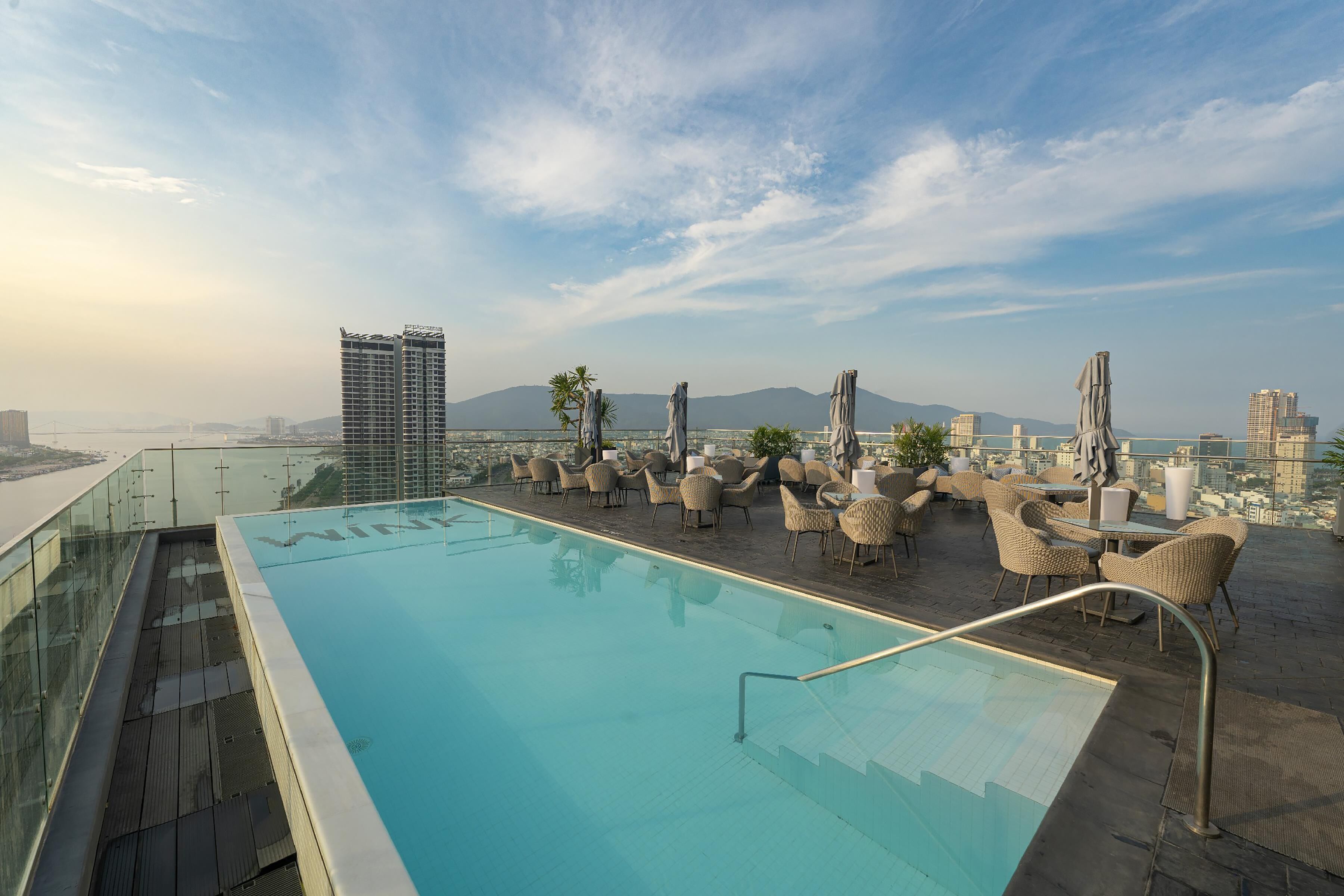 Una piscina en la azotea con paredes de vidrio transparente, mobiliario de mimbre y vistas a la ciudad.