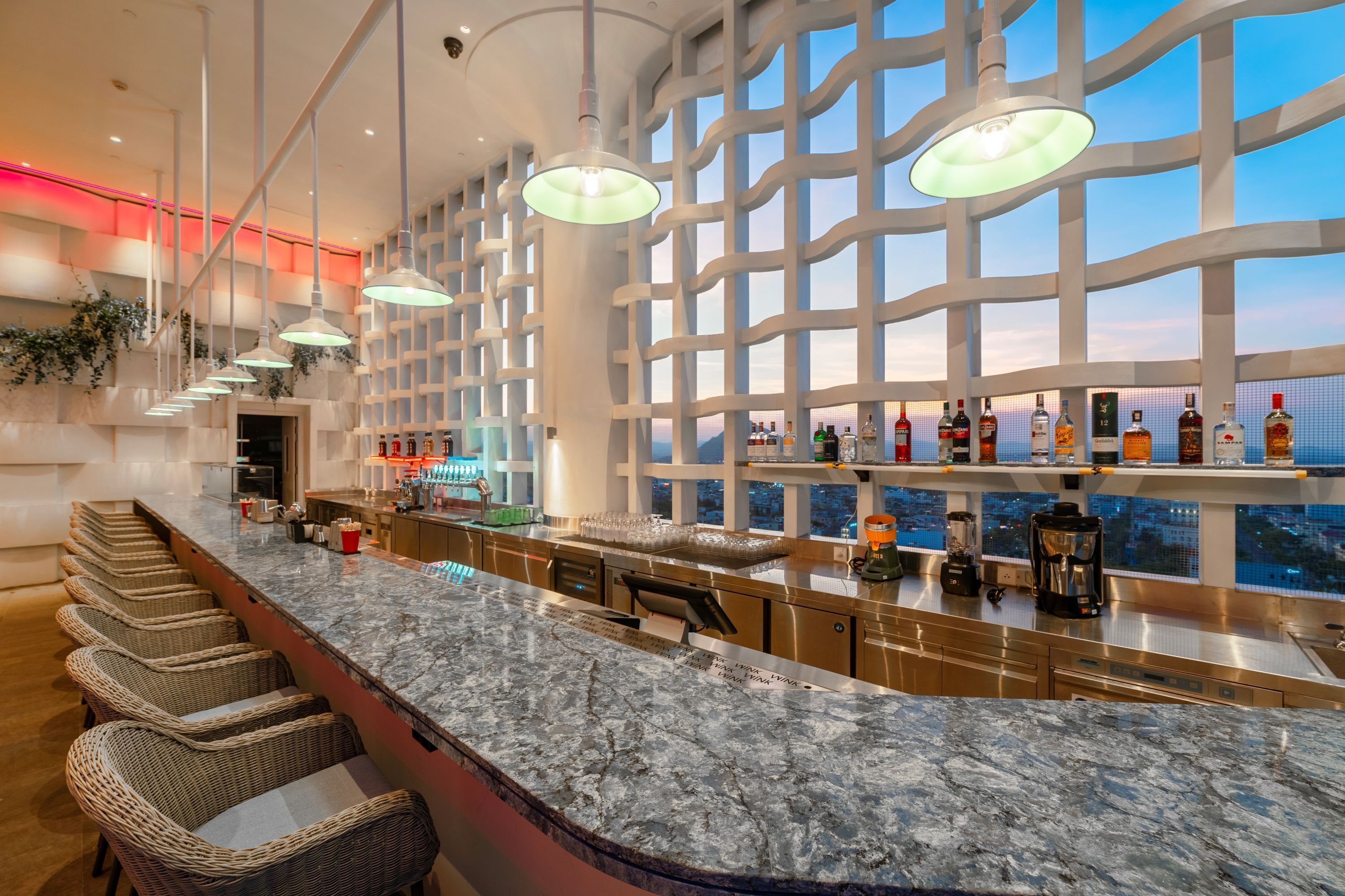 A modern bar area with a marble countertop, wicker chairs, and a view of the city through large windows.