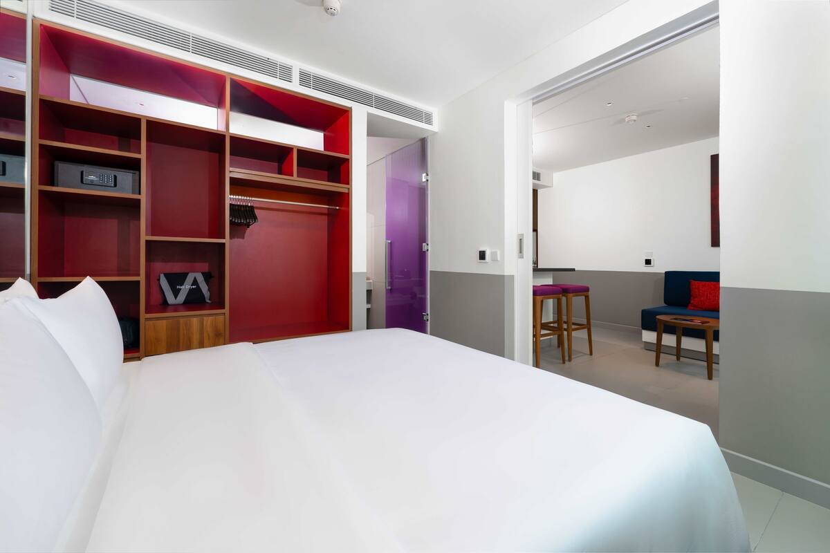 A hotel room with a large bed, a wardrobe with multiple compartments, a small seating area with a chair and stool, and a door leading to another room.