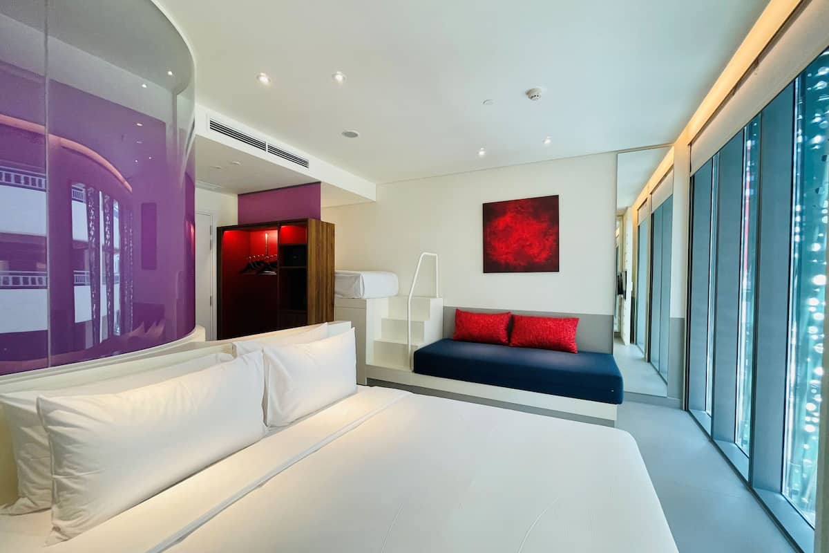 A modern hotel room with a large bed, a seating area, and a view through large windows.