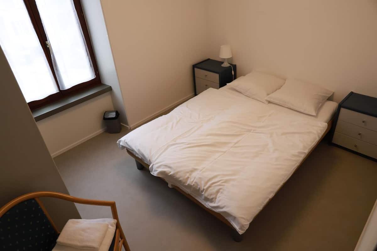 Een slaapkamer met een enkel bed, twee nachtkastjes met lampen, een stoel en een raam.