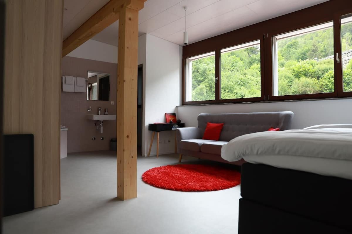 Een moderne slaapkamer met een bed, een rood tapijt, een grijze bank en een raam met uitzicht op groen.