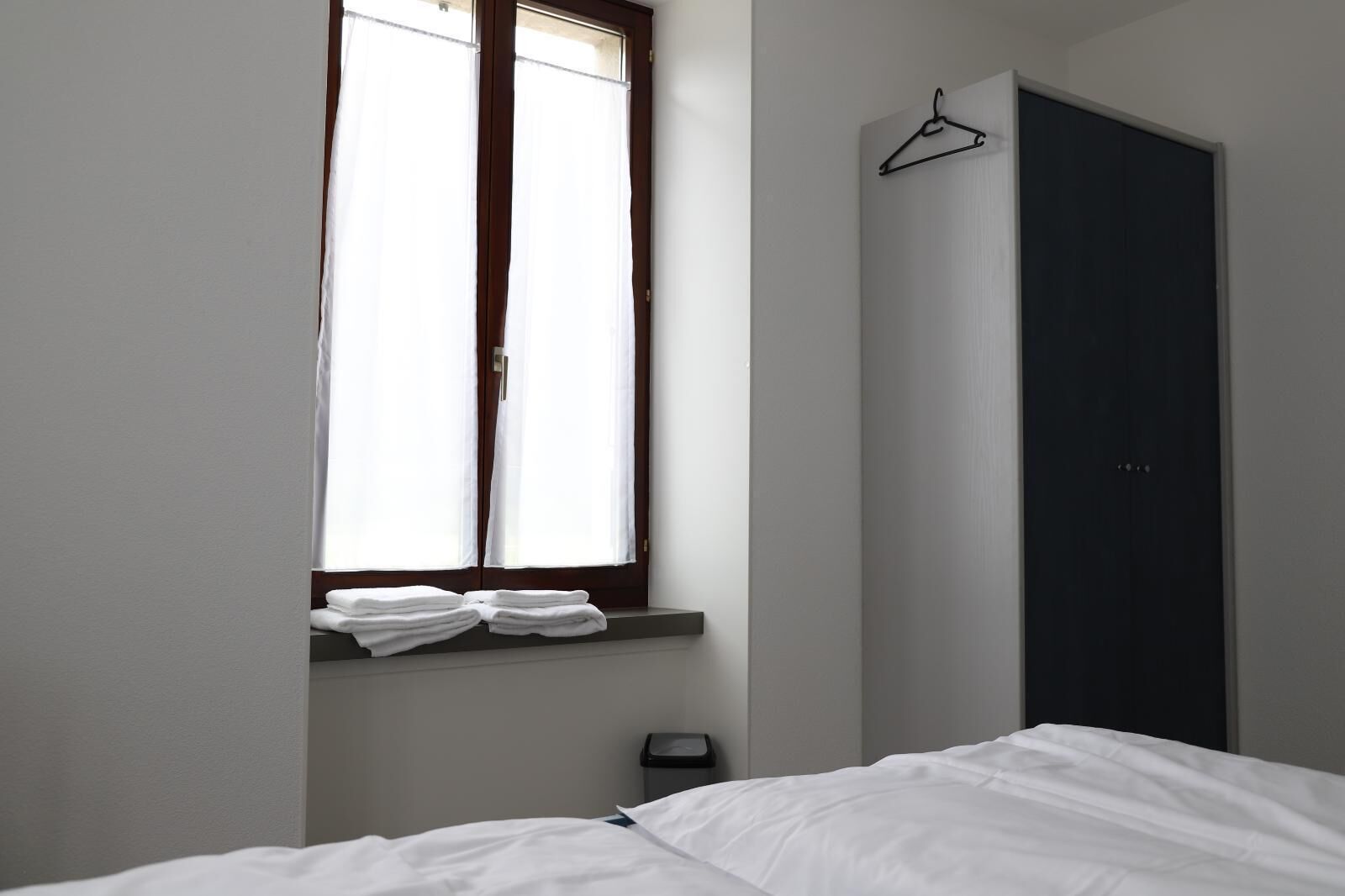 Une chambre à coucher avec un lit, une fenêtre avec des rideaux, une porte sombre et une petite étagère avec des serviettes pliées.