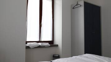 Une chambre à coucher avec un lit, une fenêtre avec des rideaux, une porte sombre et une petite étagère avec des serviettes pliées.
