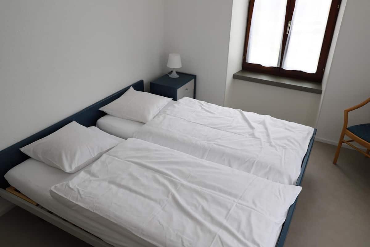 Een tweepersoonsbed met witte lakens, een nachtkastje met een lamp en een raam met gordijnen.