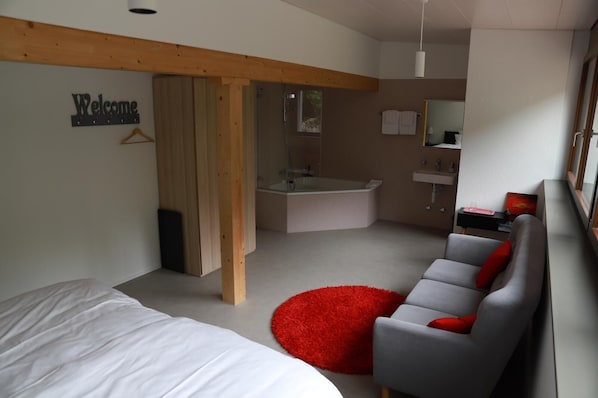 Un dormitorio moderno con una cama grande, un sofá gris con cojines rojos, un tapete suave rojo, techo de madera y un baño con bañera y lavamanos.
