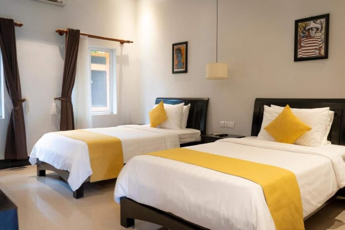 A hotel room with two beds, each with a white bedspread and a yellow accent. There is a window with curtains, a painting on the wall, and a lamp.