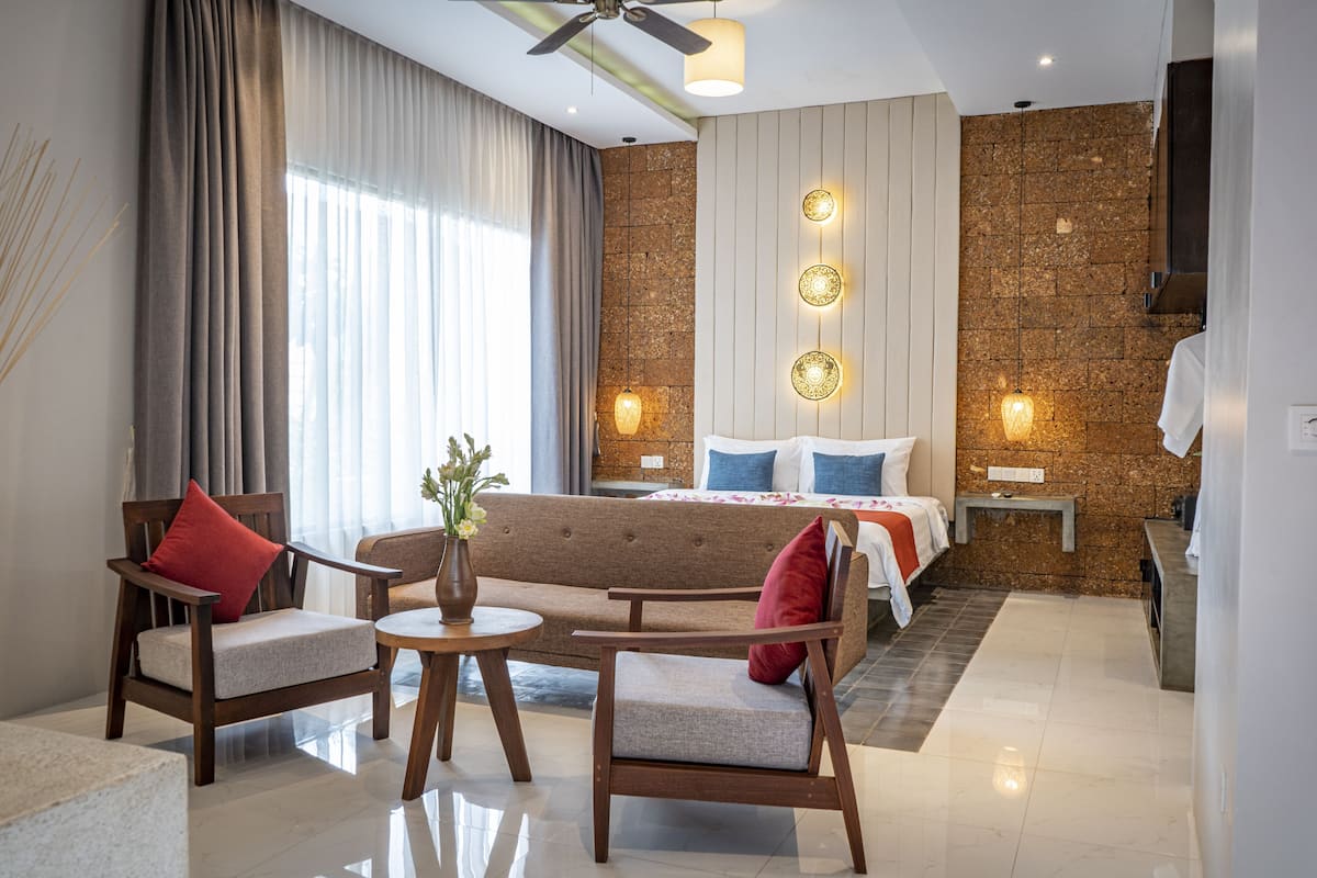 A modern hotel room with a sofa, chairs, a small table, a bed with a headboard, a stone accent wall, and a ceiling fan.