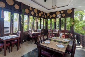 Un restaurante con mesas y sillas de madera, decoraciones de pared tejidas y ventanas amplias con follaje afuera.