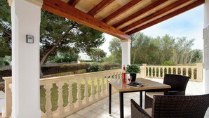Eine überdachte Terrasse mit Tisch und Stühlen, einem Blumentopf und Blick auf Bäume sowie ein Gebäude im Hintergrund.