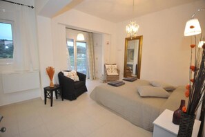 Une chambre à coucher avec un grand lit, une chaise, une petite table, un miroir et une fenêtre avec des rideaux.
