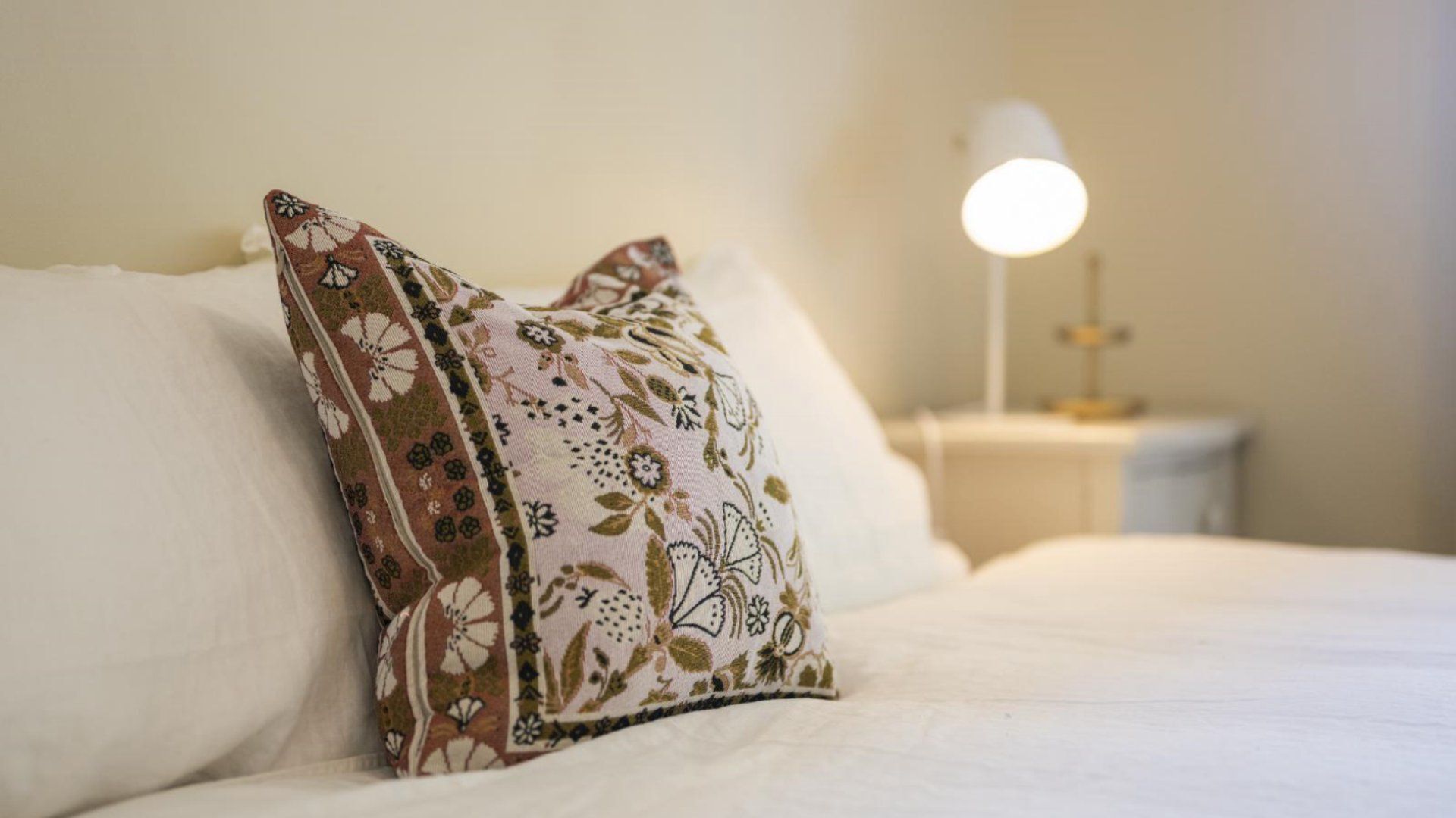 A neatly made bed with white linens and a decorative pillow with a floral pattern.