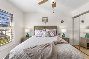 A bedroom with a bed, two nightstands, a ceiling fan, a window with blinds, and a mirror.
