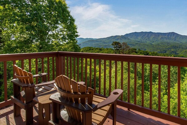 Terraza de madera con sillas Adirondack y una mesa, con vistas a un paisaje montañoso.