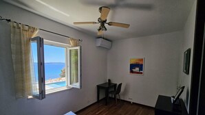 A room with a bed, a ceiling fan, a window with a view of the sea, a small table with a chair, and a painting on the wall.