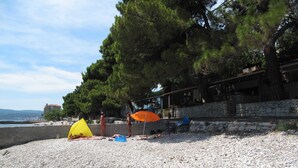 A pebbled beach with tents and umbrellas, a stone wall, and a building with a balcony overlooking the sea.