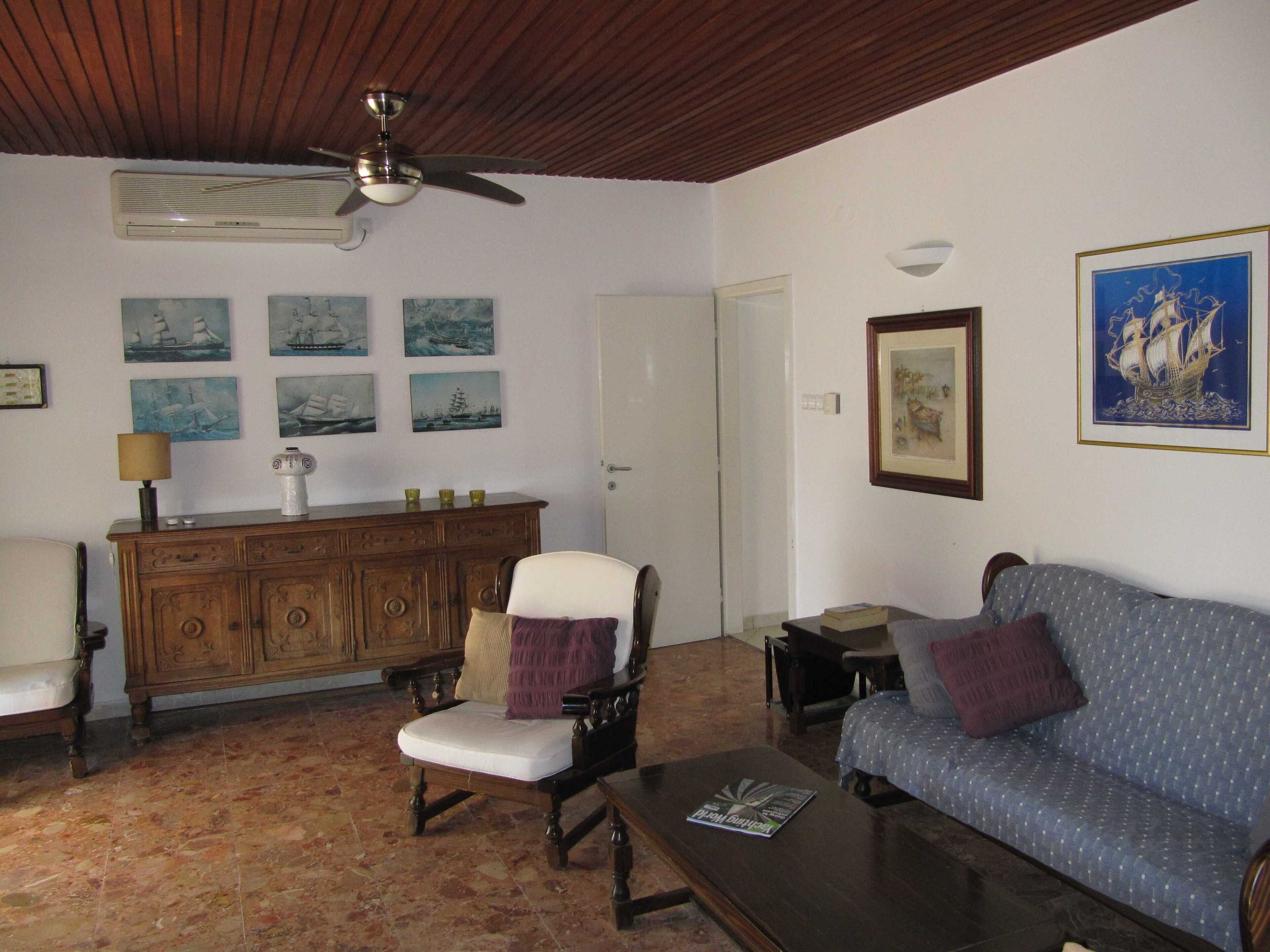 A living room with a wooden ceiling, a ceiling fan, a white sofa, a blue sofa, a wooden cabinet, and a chair.