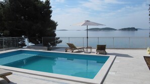 A rectangular swimming pool with a white border, surrounded by a tiled deck. There are lounge chairs and a single umbrella providing shade. A metal railing encloses the deck, and a body of water with distant landforms is visible in the background.