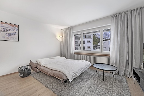 Ein Schlafzimmer mit einem Bett, einem kleinen runden Tisch, einem Heizkörper und einem Fenster mit Vorhängen.