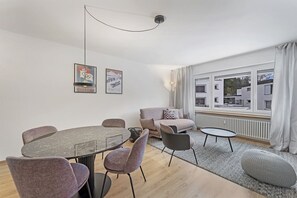 Ein modernes Wohnzimmer mit einem runden Esstisch, mehreren Stühlen, einer Couch und einem großen Fenster mit Vorhängen.